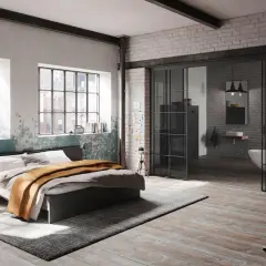 Ein gemütliches **Bett unter Fenster** mit industriellem Charme. Daneben ein offener Badbereich mit Wanne.